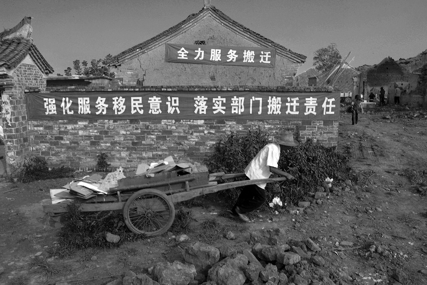 Under banners that read “Wholeheartedly Support Relocation” and “Strengthen Your Awareness to Serve Migrants to Fulfill Relevant Departments’ Responsibilities in Relocation,” a farmer pulls his belongings in a handcart to load them on a truck to transport them to his new home.