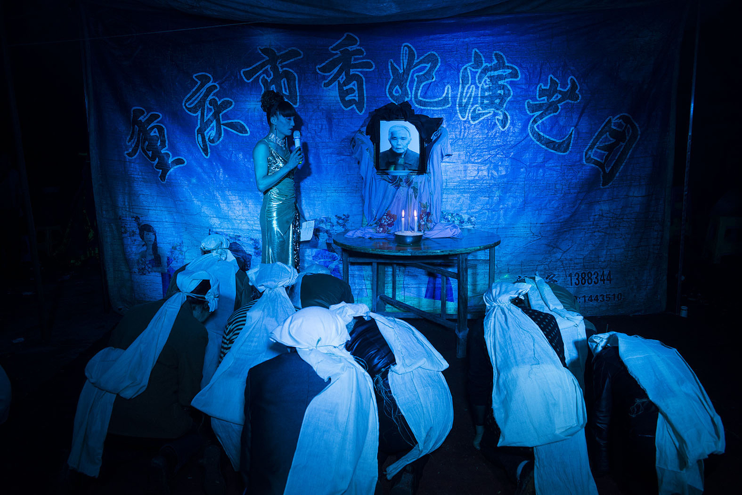 Family members kneel in front of a portrait of the departed as Liangzi hosts a funeral ceremony, April 10, 2014. As a funeral performer, Liangzi is skilled at singing, dancing, and wailing. She is proud of her wailing skill, and she tries to cry naturally at every funeral.