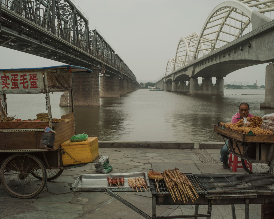 A food vendor awaits customers on the bank of the Songhua River in Harbin, beneath bridges used by Chinese Eastern Railway. Chinese in name alone, the railway was built on a strip of land controlled by Russia that was exempt from the jurisdiction of local Chinese law in a deal brokered between the two countries after China lost the Sino-Japanese war. The old bridge, seen at left, was built by the Russians at the beginning of the 20th century; the newer one was built by the Chinese.